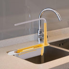 A grey telescopic sink storage rack shown fully extended over a kitchen sink, holding a sponge and brush.